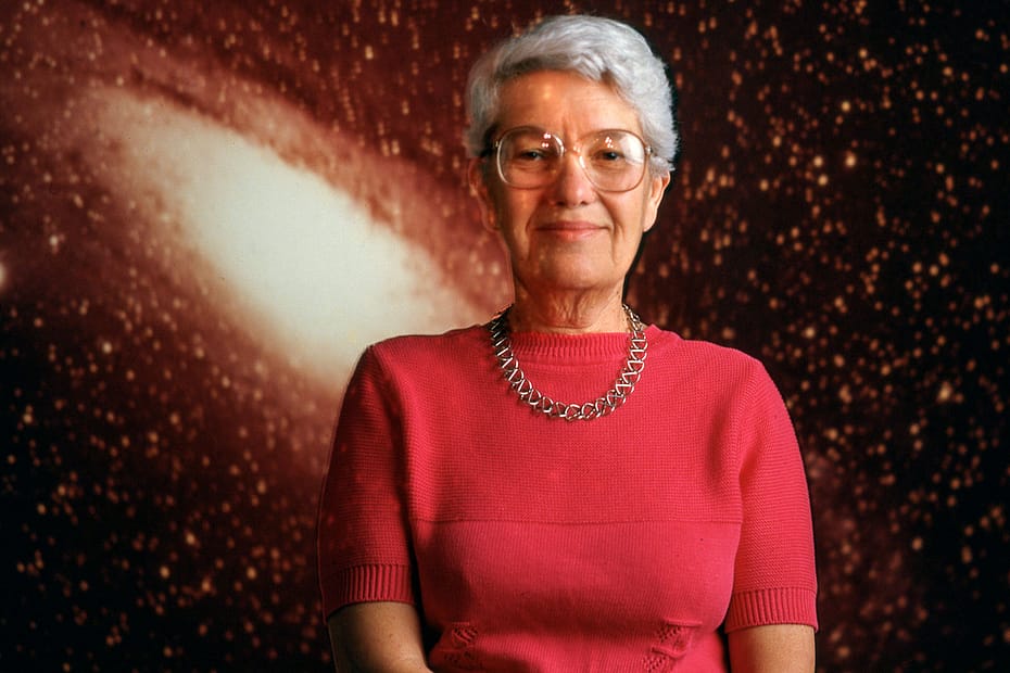 vera Rubin usando uma roupa vermelha, com cabelos grisalhos e curtos, e óculos. Ela usa um colar no pescoço, transmitindo um ar de sofisticação. Ao fundo, uma galáxia em tons alaranjados, ressaltando sua conexão com o campo da astronomia e a vastidão do universo que ela ajudou a explorar.
