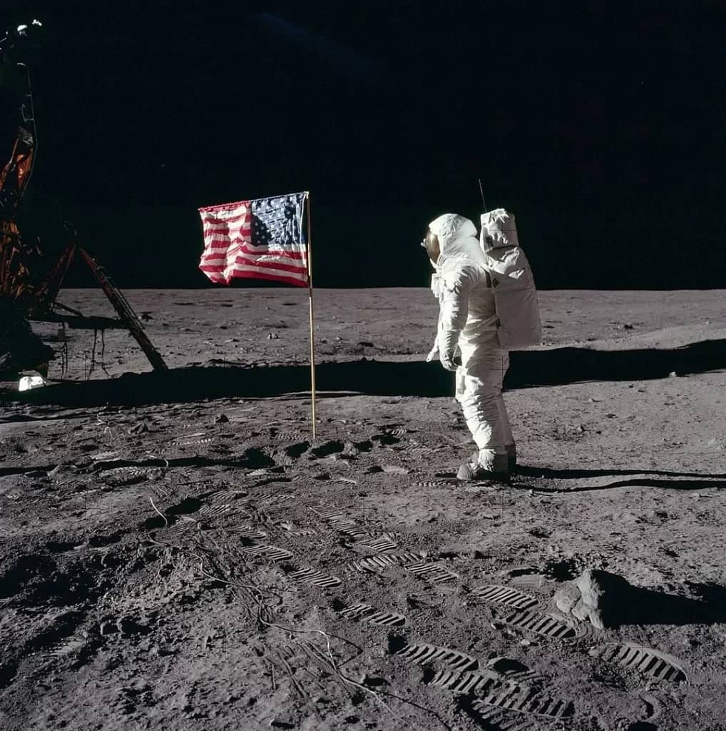 Apollo 11 Buzz Aldrin, o astronauta da missão Apollo 11, saudando a bandeira dos Estados Unidos na superfície da Lua em 1969. Ele está vestido com seu traje espacial, com o visor refletindo a paisagem lunar ao fundo, enquanto a bandeira americana ondula ao vento (apesar da falta de atmosfera) em um gesto simbólico de conquista.