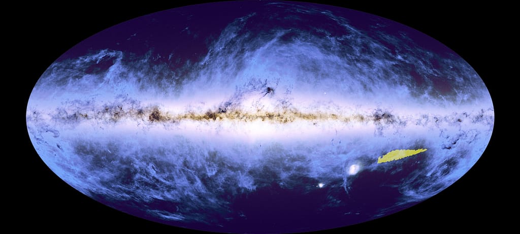 Uma imagem oval azul escura, com uma faixa brilhante que se estende da esquerda para a direita, de onde nuvens azul-claras parecem se espalhar pelo restante da forma. Essa faixa brilhante é interrompida por estruturas mais escuras, semelhantes a nuvens. No canto inferior direito da oval, há uma pequena área destacada em amarelo brilhante, indicando a localização do mosaico completo no contexto deste mapa global da nossa galáxia, a Via Láctea.