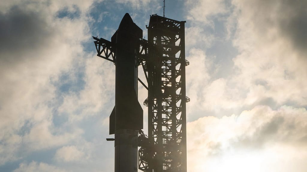 Imagem da Starship da SpaceX antes do sexto teste de voo, posicionada na plataforma de lançamento. O foguete está montado em sua estrutura de lançamento, com os motores e sistemas de propulsão visíveis. O céu claro ou parcialmente nublado no fundo destaca a imponente presença do foguete, que se prepara para sua missão espacial. Detalhes dos suportes e da infraestrutura de lançamento complementam a cena, mostrando a preparação meticulosa para o voo