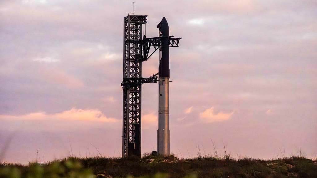 Imagem do foguete spacex Ship completa no pad de lançamento, iluminada por um céu laranja ao fundo, pronta para decolar. O foguete da SpaceX, projetado para missões espaciais interplanetárias, está em preparação para novas aventuras, incluindo viagens à Lua, Marte e além