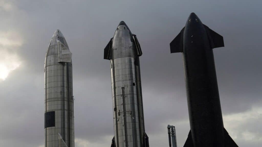 três naves Starship aguardando montagem na base de lançamento Starbase, em um dia nublado. As naves, projetadas pela SpaceX para missões espaciais ambiciosas, estão alinhadas enquanto se preparam para futuras decolagens rumo à Lua, Marte e além.