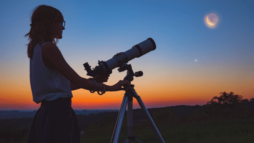 Imagem de um astrônomo observando o céu com um telescópio, enquanto o céu mostra o amanhecer e é possível ver a lua e Vênus brilhando, no contexto de uma noite clara com a aurora ao fundo.
