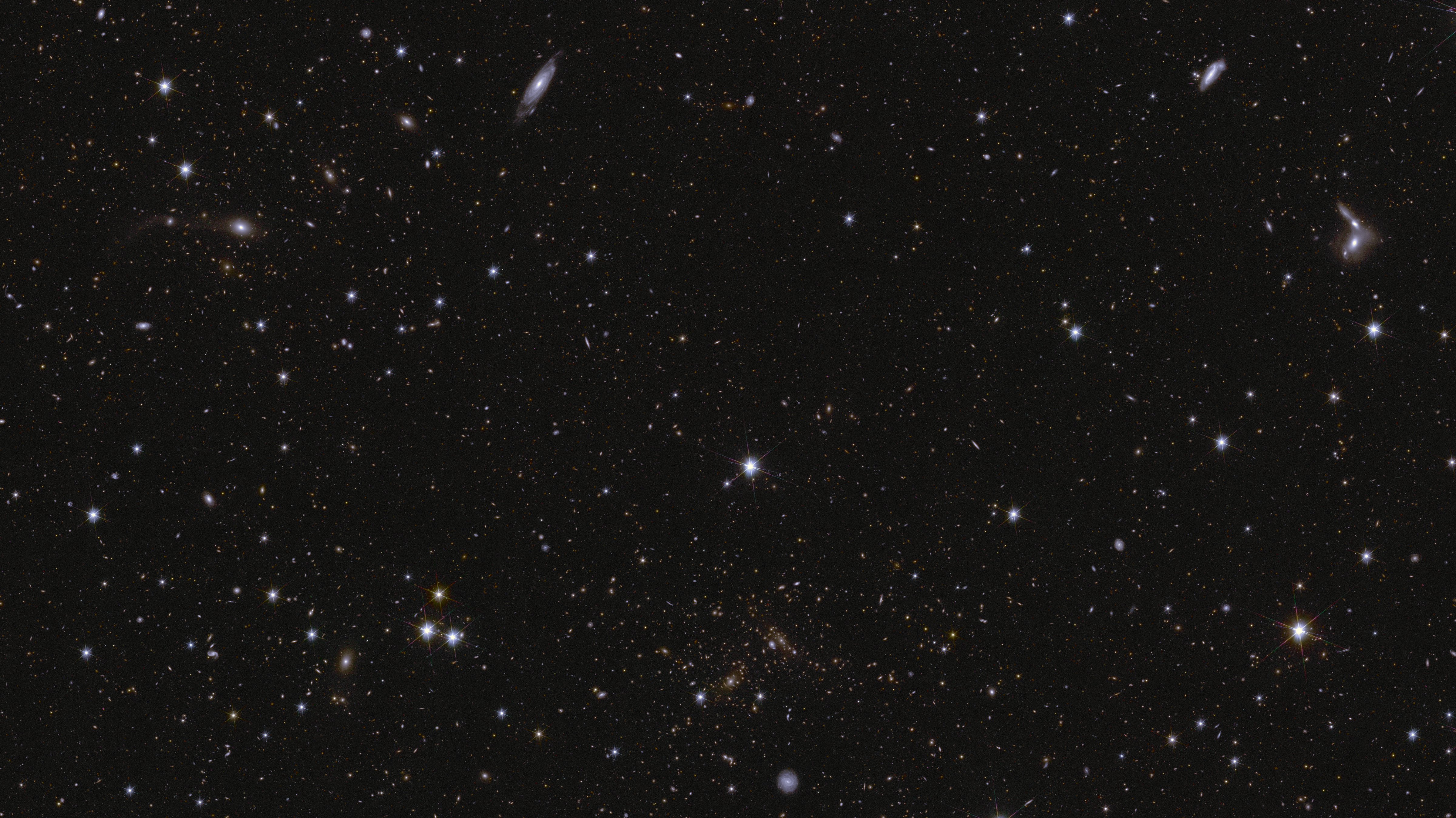 Um mar de estrelas e galáxias brilha contra um fundo preto. Várias galáxias podem ser identificadas por suas formas alongadas e/ou braços espirais. Algumas galáxias são vistas de perfil, enquanto uma galáxia espiral proeminente no centro inferior da imagem aparece de frente. No canto direito, entre o meio e a parte superior da imagem, estão algumas galáxias em interação. Também são visíveis aglomerados de galáxias, especialmente próximos ao centro inferior, onde estruturas distorcidas em forma de arco representam lentes gravitacionais. As estrelas mais brilhantes da imagem apresentam picos de difração.