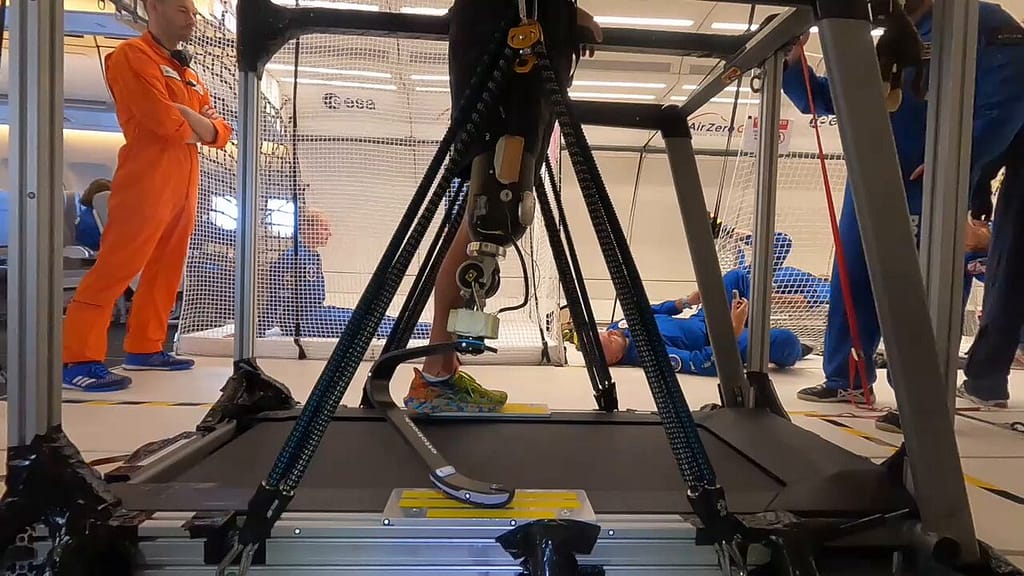 "Close na prótese de perna de John McFall enquanto ele corre na esteira durante um voo parabólico. A prótese é visível em detalhes, com foco na adaptação tecnológica para ambientes de microgravidade. Ao fundo, outros astronautas, vestidos com trajes espaciais azuis, flutuam enquanto observam o experimento, criando um contraste com a cena de teste da prótese em movimento.