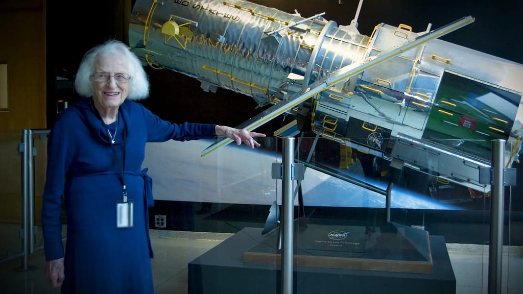 magem de Nancy Grace Roman, astrônoma conhecida como "Mãe do Hubble", já idosa, sorrindo em frente a uma maquete do Telescópio Espacial Hubble.
