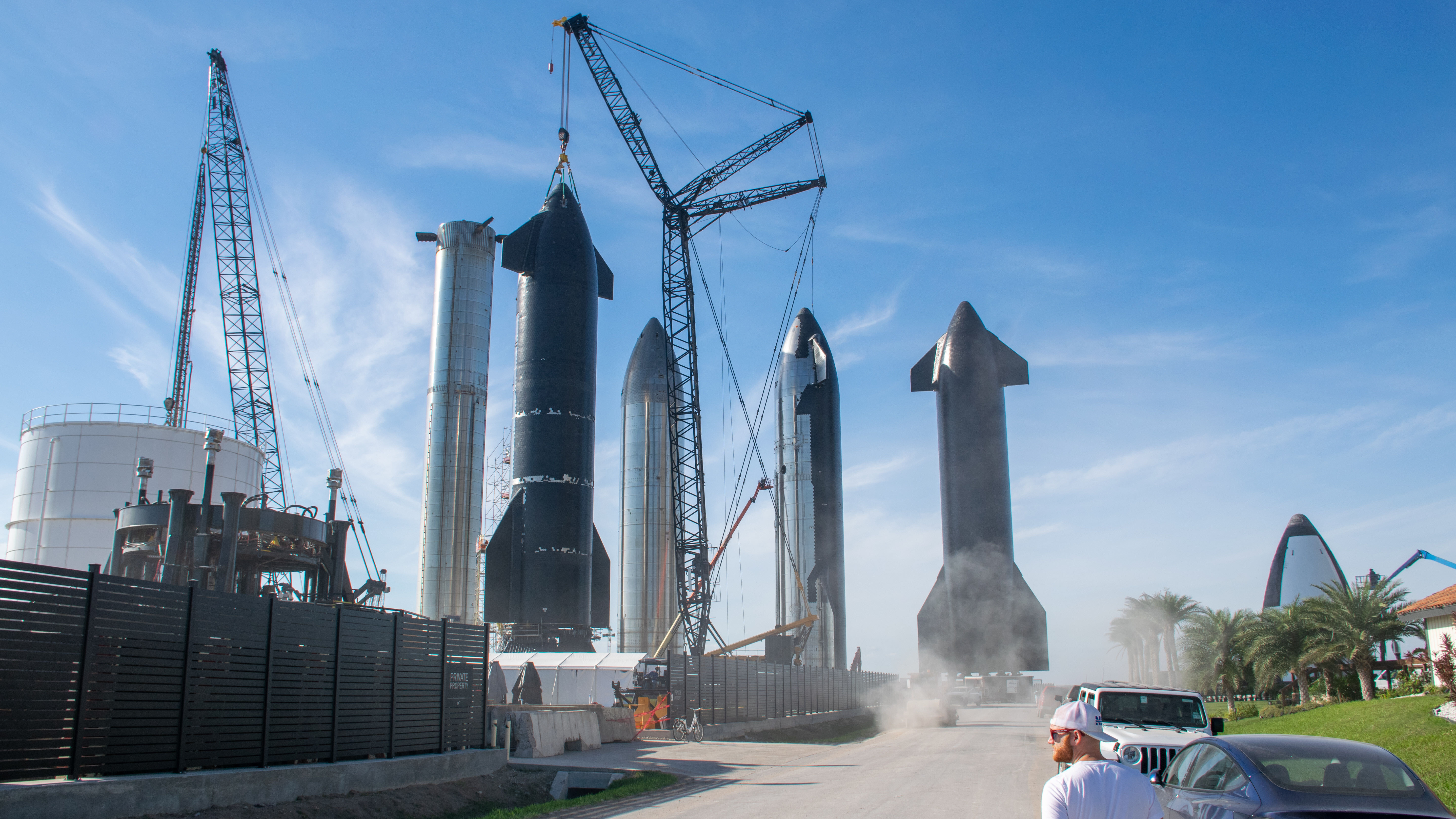 Visão ampla da Starbase da SpaceX com quatro naves Starship sendo transportadas por guindastes e um booster Super Heavy ao fundo.