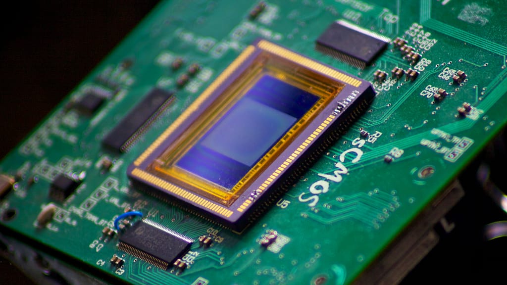 O sensor de imagem CMOS, uma das Tecnologias Desenvolvidas pela Astronomia, é montado em uma placa verde característica, com detalhes de contatos dourados visíveis. A placa verde, composta por circuitos intricados, e os contatos dourados destacam-se por seu brilho metálico, facilitando a transmissão de sinais elétricos para o processamento de imagens. O sensor CMOS captura a luz e converte-a em imagem, utilizando fotodetectores e transistores, uma inovação que tem impacto em diversas áreas além da exploração espacial.