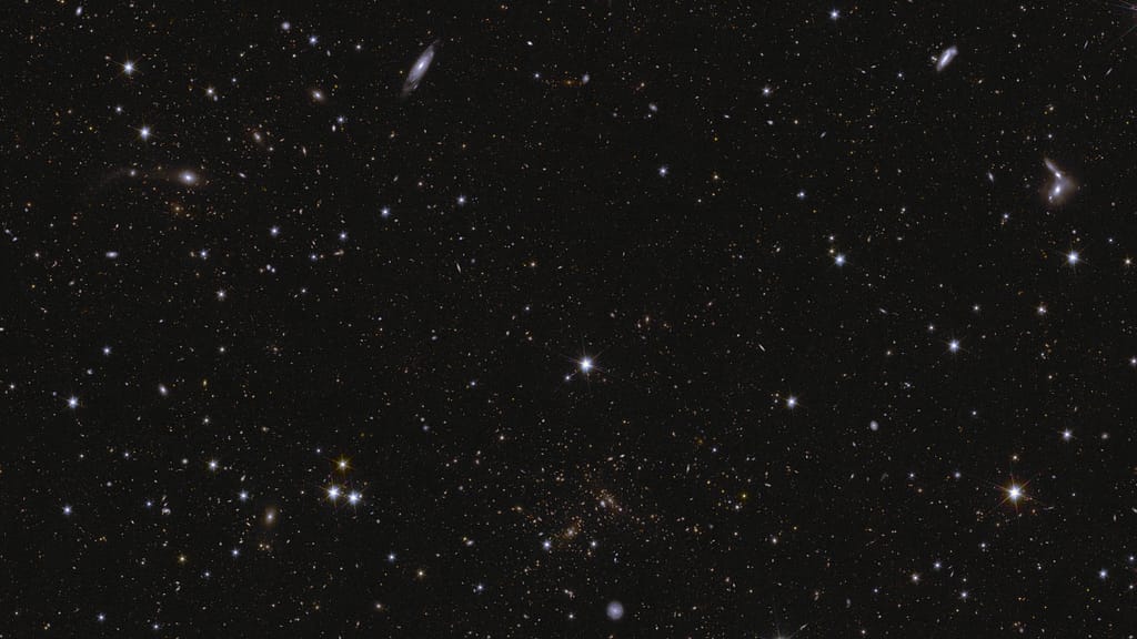 Um mar de estrelas e galáxias brilha contra um fundo preto. Várias galáxias podem ser identificadas por suas formas alongadas e/ou braços espirais. Algumas galáxias são vistas de perfil, enquanto uma galáxia espiral proeminente no centro inferior da imagem aparece de frente. No canto direito, entre o meio e a parte superior da imagem, estão algumas galáxias em interação. Também são visíveis aglomerados de galáxias, especialmente próximos ao centro inferior, onde estruturas distorcidas em forma de arco representam lentes gravitacionais. As estrelas mais brilhantes da imagem apresentam picos de difração.
