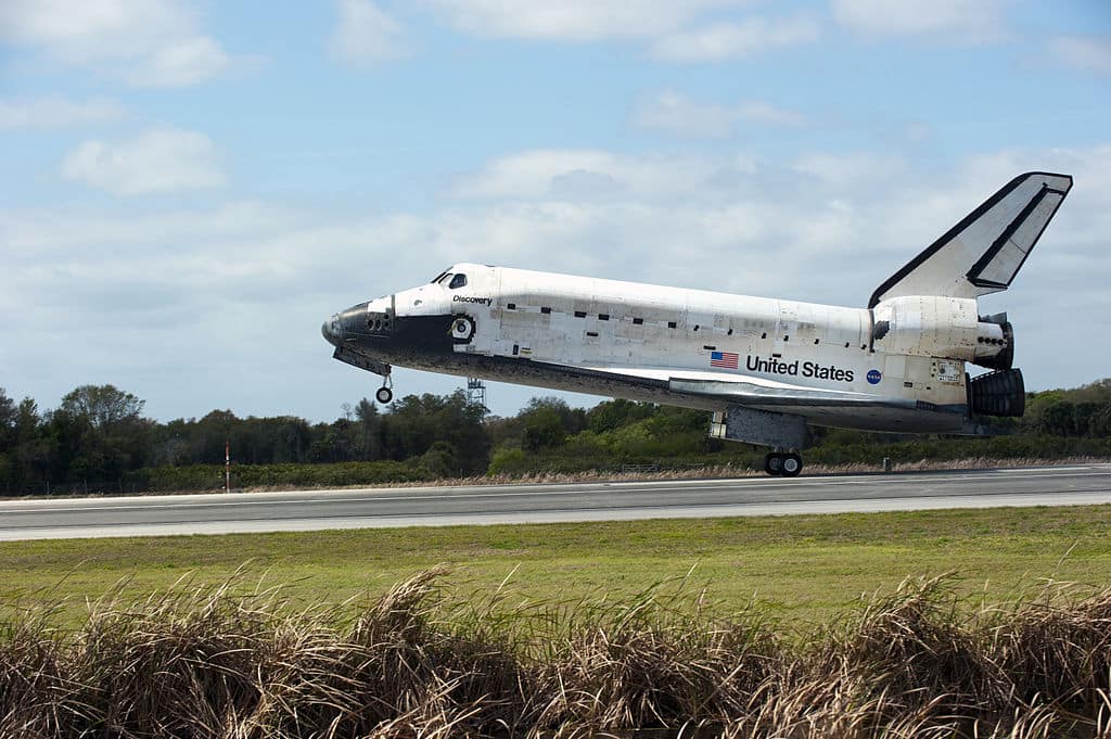 Ônibus espacial Discovery pousando na Terra durante sua última missão, STS-133, em 9 de março de 2011, com fumaça visível nas rodas tocando a pista após o contato, marcando o fim de sua trajetória histórica.
