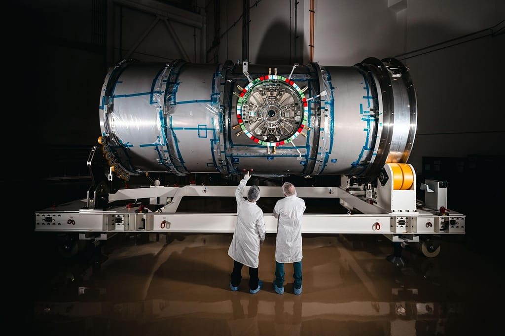 Dois técnicos em trajes de sala limpa estão em frente ao HALO (Habitation and Logistics Outpost) na instalação da Northrop Grumman em Gilbert, Arizona. O módulo cilíndrico metálico, recentemente desembrulhado, está repousando sobre um suporte branco. Um dos técnicos aponta para a parte frontal do módulo. Tecnologias do Programa Artemis da NASA