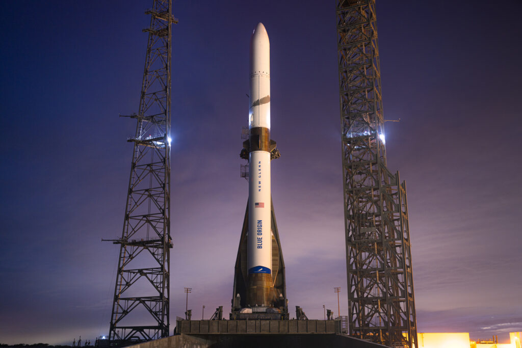 Visão próxima do foguete New Glenn ao entardecer, destacando sua estrutura branca com a bandeira dos EUA, o nome 'Blue Origin' e 'New Glenn' escritos em azul. A iluminação suave do pôr do sol realça os detalhes do foguete antes do lançamento