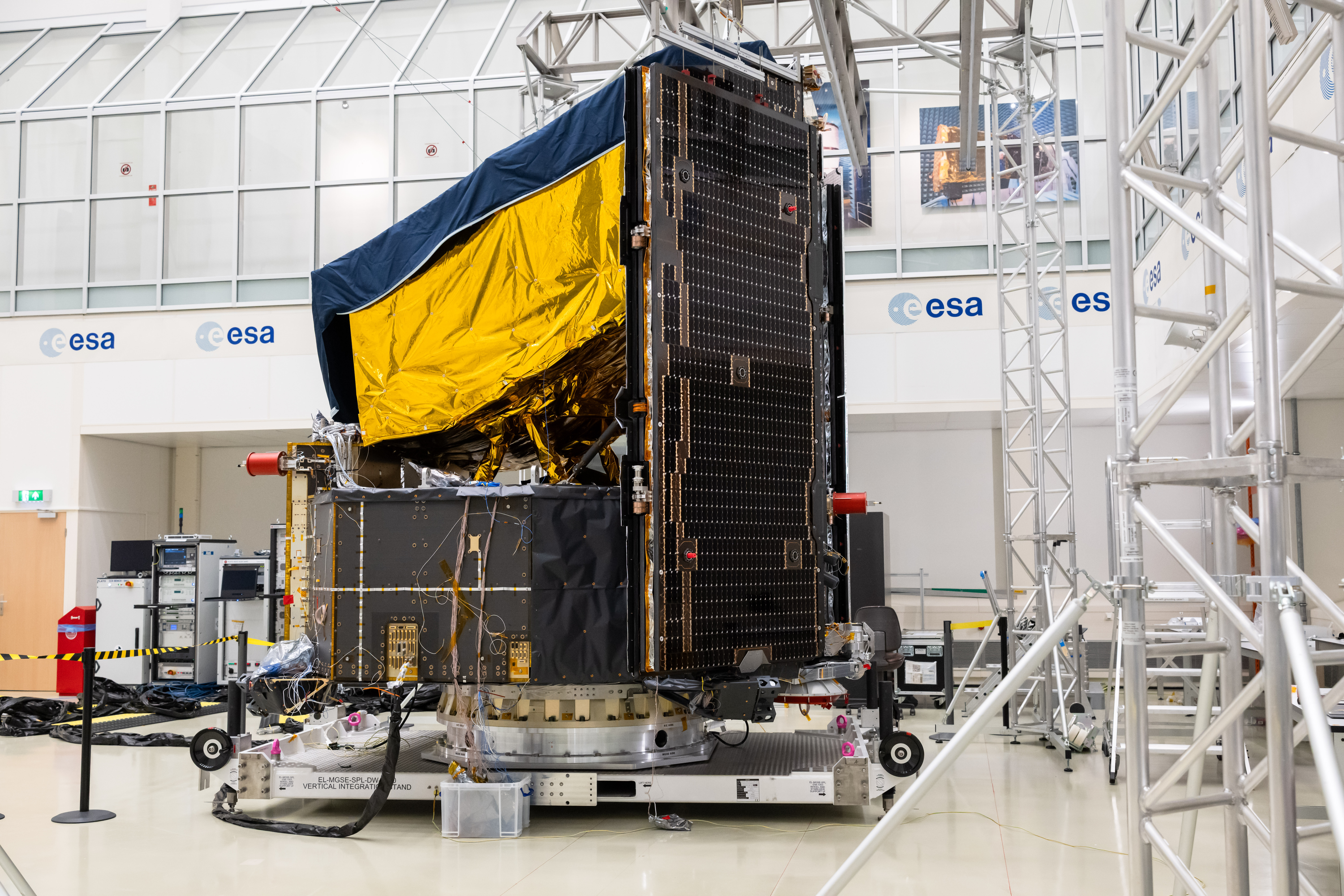 Um grande satélite está posicionado dentro de uma sala limpa intensamente iluminada. As laterais da parte superior do satélite estão envoltas em um revestimento dourado (mantas térmicas), enquanto o topo está coberto por uma manta azul. A parte inferior consiste em uma estrutura preta de formato retangular. A espaçonave está montada sobre uma plataforma com rodas; vários instrumentos e andaimes são visíveis em primeiro plano.