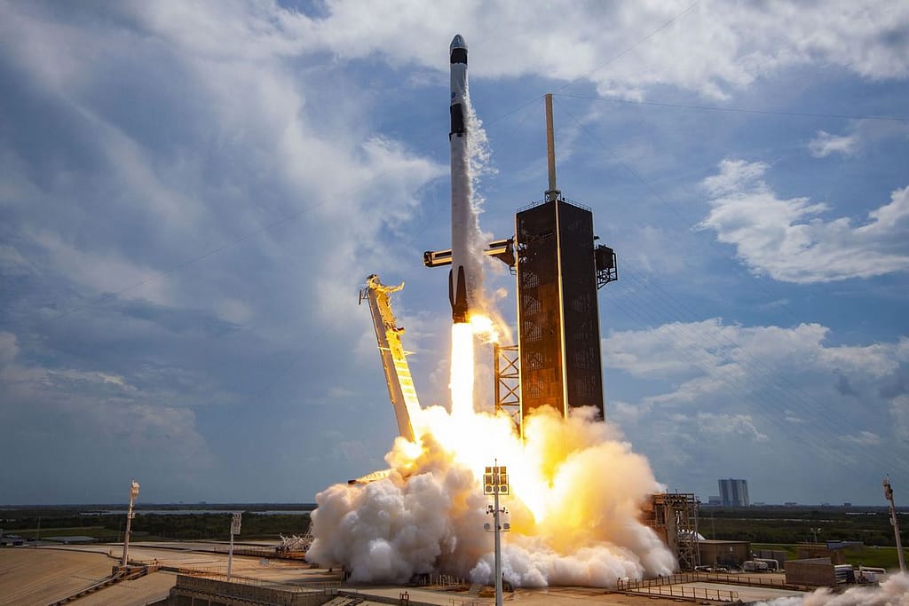 Foguete Falcon 9 sendo lançado da base, com fumaça e chamas saindo dos motores enquanto sobe em direção ao céu azul.