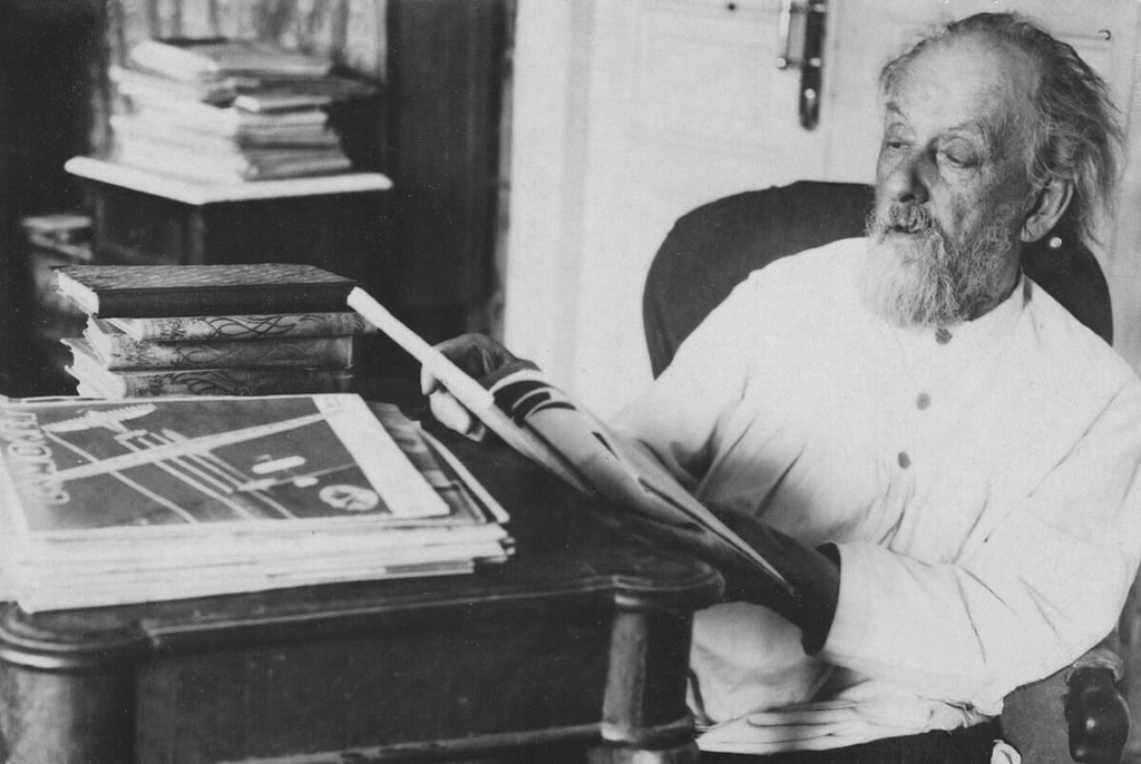 Konstantin Tsiolkovski está em uma sala, concentrado enquanto lê um livro. Com sua longa barba branca e óculos redondos, ele aparenta estar imerso em seus estudos. O ambiente ao redor sugere um espaço de trabalho, refletindo sua dedicação às teorias que revolucionaram a astronáutica.