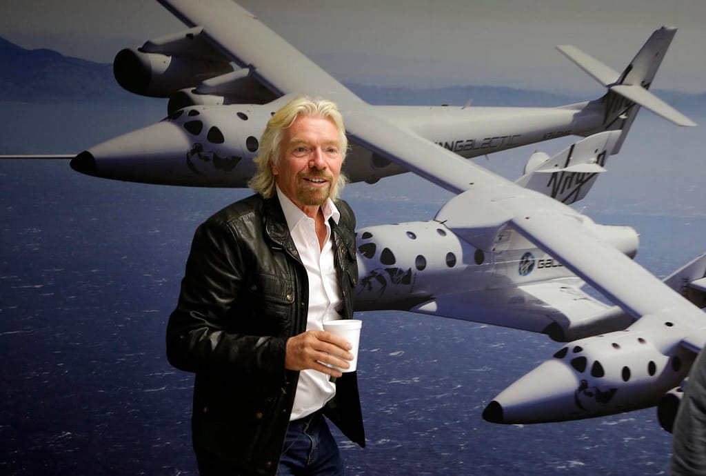 Richard Branson dando uma palestra, vestindo uma jaqueta preta. Ao fundo, há um banner na parede com a imagem e o nome da nave SS Unity, destacando o contexto aeroespacial do evento.