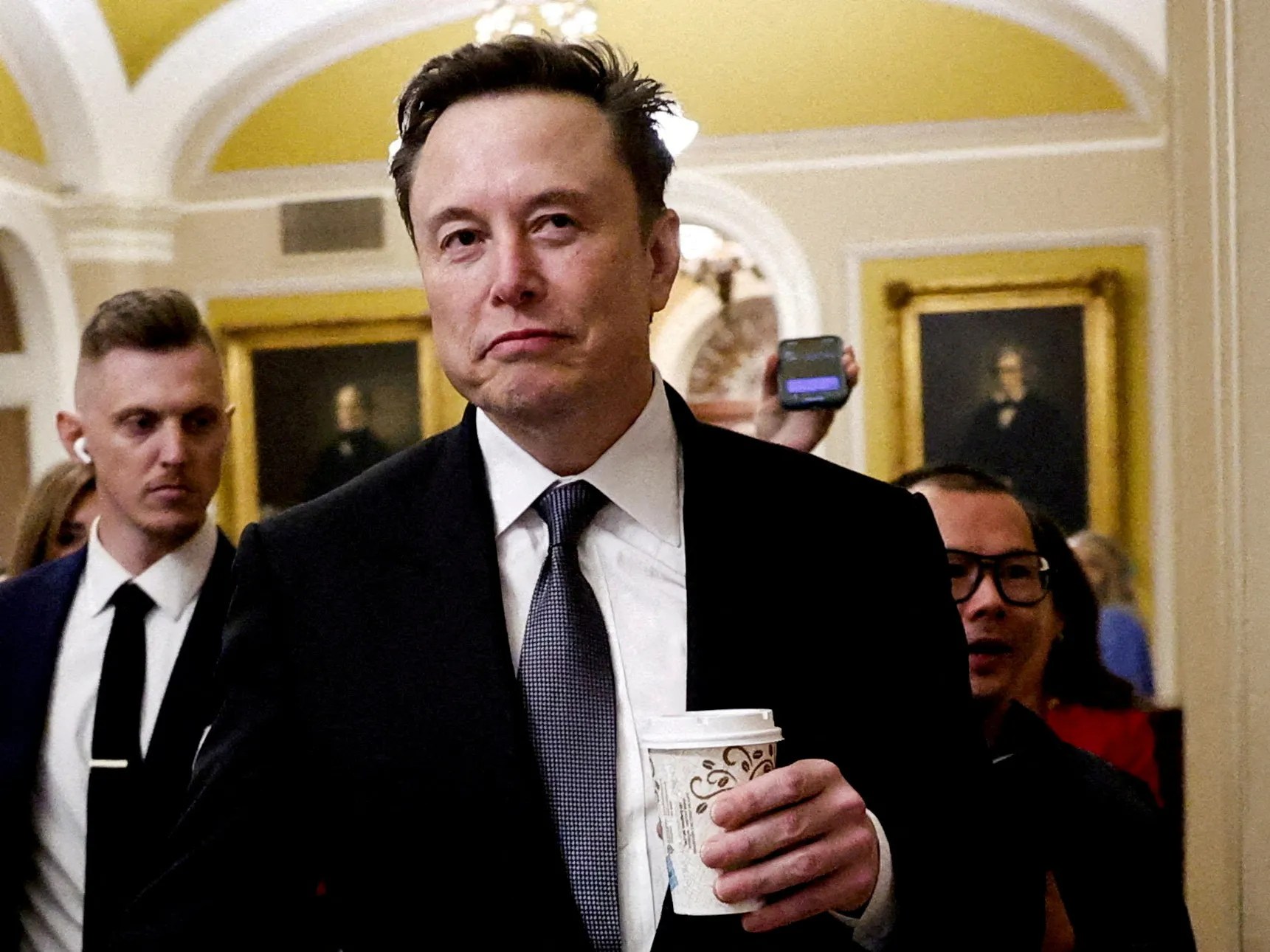 Elon Musk usando terno em um evento, segurando um copo de café e olhando para o lado com expressão pensativa.