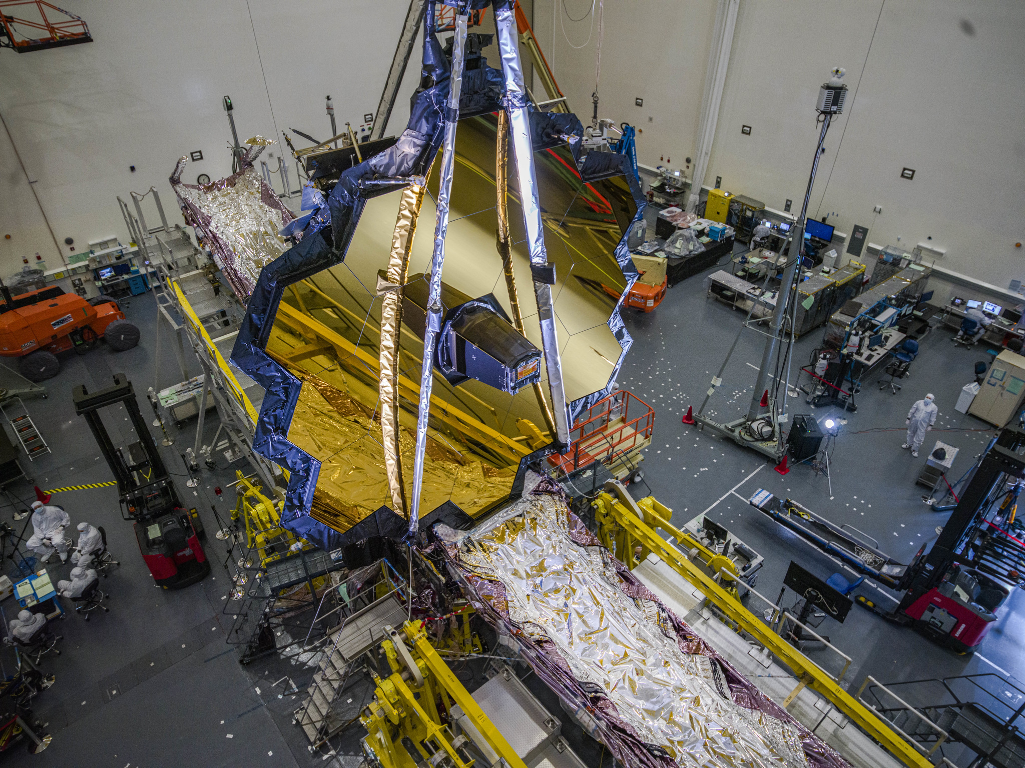 Um Telescópio Webb totalmente montado, apoiado por estruturas de suporte, ocupa o espaço dentro da sala limpa da Northrop Grumman. No centro da imagem está o espelho hexagonal revestido de ouro do telescópio, que é dobrado para trás em ambos os lados. Abaixo do espelho está a estrutura do protetor solar. O protetor solar é composto por cinco finas camadas em forma de pipa, cada uma construída com um material que se parece muito com folha de prata. Três técnicos, cada um usando equipamento branco com controle de contaminação, podem ser vistos em guindastes laranja ao redor do telescópio. Um técnico está em primeiro plano, um no meio do plano e um no fundo. Uma linha azul-escura percorre o topo das paredes brancas da sala. O teto da sala também é branco.