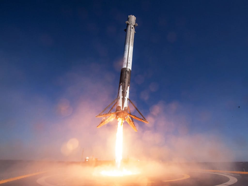 Foguete Falcon 9 realizando seu primeiro pouso bem-sucedido em uma balsa no oceano, com fumaça ao redor e estrutura metálica da balsa visível sob o céu limpo.