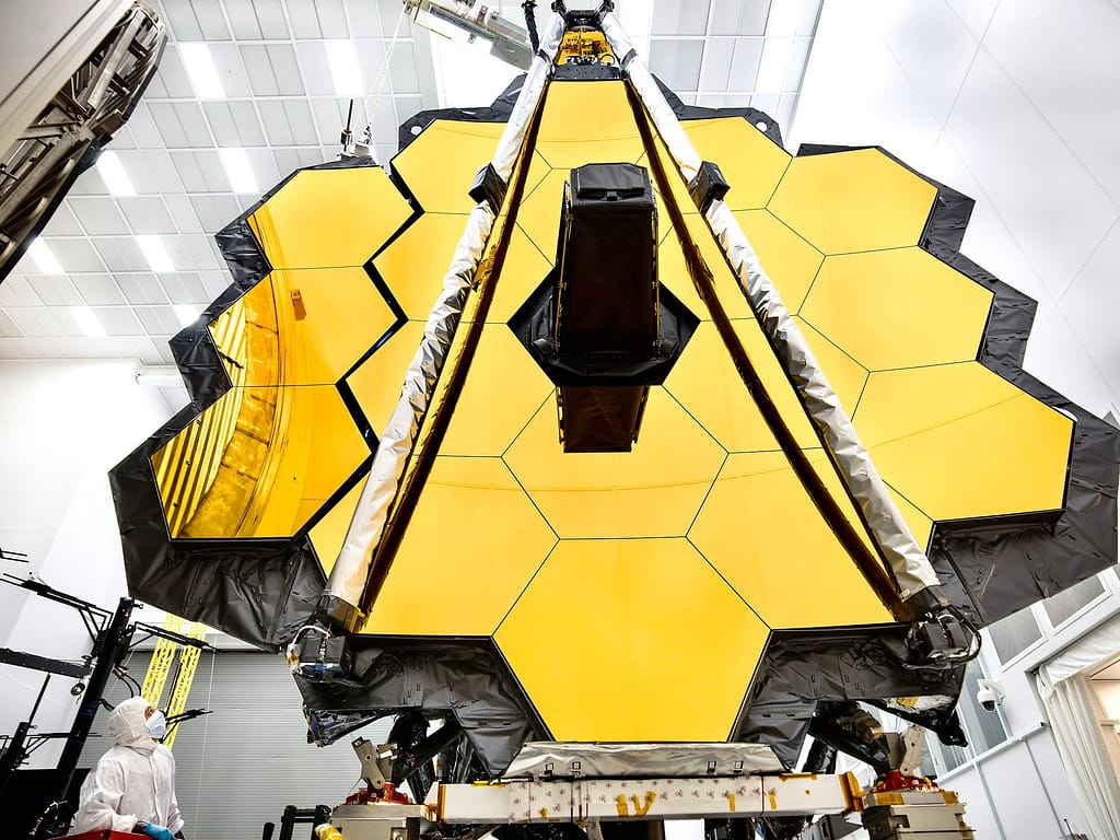 O imponente espelho primário do Telescópio Espacial James Webb da NASA está dentro de uma sala limpa no Centro Espacial Johnson da NASA, em Houston, onde passará por seu último teste criogênico antes de ser lançado ao espaço em 2018. Em preparação para o teste, as "asas" do espelho (que consistem em três segmentos de cada lado) foram abertas. Esta foto mostra uma asa totalmente desdobrada e outra que está a momentos de se desdobrar completamente. Um engenheiro observa o movimento. O Telescópio Espacial James Webb é o observatório espacial mais avançado do mundo. Esta maravilha da engenharia foi projetada para desvendar alguns dos maiores mistérios do universo, desde a descoberta das primeiras estrelas e galáxias que se formaram após o Big Bang até o estudo das atmosferas de planetas ao redor de outras estrelas. É um projeto conjunto da NASA, da ESA (Agência Espacial Europeia) e da Agência Espacial Canadense.