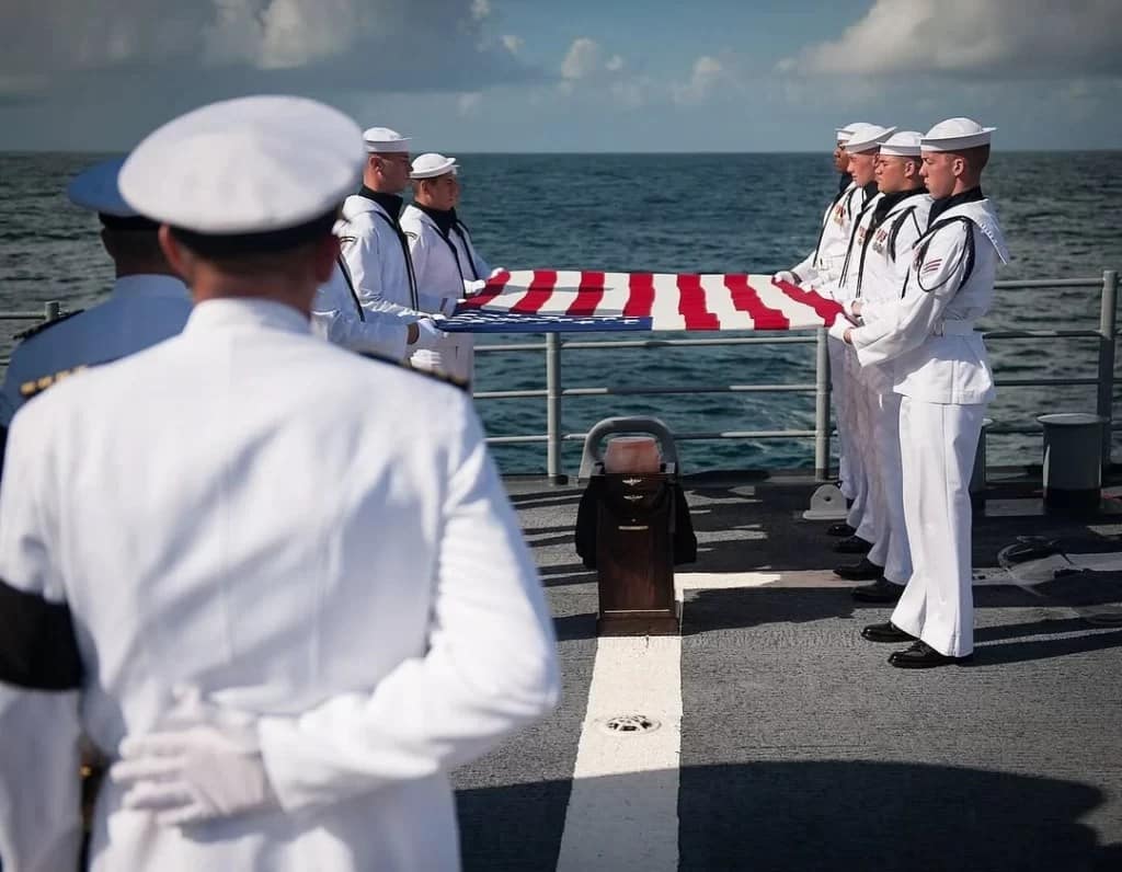 marinheiros, alinhados no alto-mar, enquanto estendem a bandeira americana. Eles estão em fila, fazendo uma saudação a Neil Armstrong e à missão Apollo 11. A bandeira está visivelmente em boas condições, com cores vibrantes, e o mar calmo ao fundo, simbolizando o respeito e homenagem à histórica conquista.