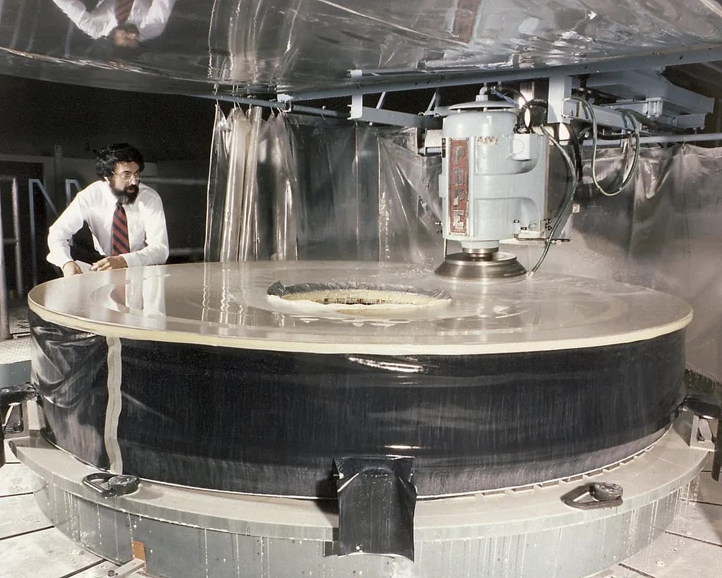 Lixamento Preliminar do Espelho Primário do Telescópio Espacial (2,4 metros) nas Instalações F-25 da Perkin Elmer, março de 1979. Esta fotografia mostra o espelho primário do Telescópio Espacial Hubble sendo lixado nas grandes instalações de fabricação de óptica da Perkin-Elmer Corporation. Após o espelho de 8 pés de diâmetro ser lixado e polido, a superfície de vidro foi revestida com uma camada reflexiva de alumínio e uma camada protetora de fluoreto de magnésio, com espessura de 0,1 e 0,025 micrômetros, respectivamente. O propósito do Hubble, o telescópio óptico mais complexo e sensível já construído, é estudar o cosmos a partir de uma órbita baixa da Terra. Ao colocar o telescópio no espaço, os astrônomos conseguem coletar dados livres da atmosfera terrestre. O Marshall Space Flight Center foi responsável pelo projeto, desenvolvimento e construção do observatório, e a Perkin-Elmer Corporation em Danbury, Connecticut, desenvolveu o sistema óptico e os sensores de orientação. Crédito da imagem: NAS