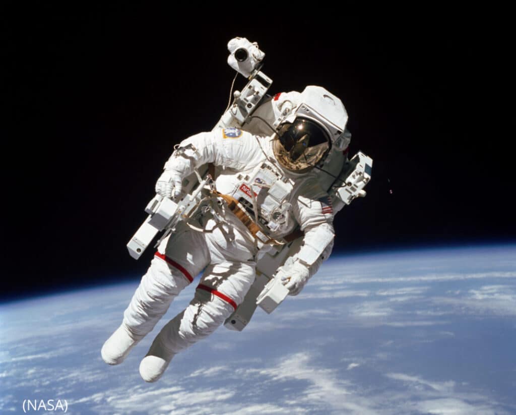Bruce McCandless II flutuando no espaço durante a missão STS-41-B do ônibus espacial Challenger, em fevereiro de 1984. Ele está usando um traje espacial, sem estar preso à nave-mãe, representando a primeira caminhada no espaço livre. Ao fundo, o vasto e escuro espaço sideral, com a Terra parcialmente visível abaixo, e as estrelas pontilhando o céu.