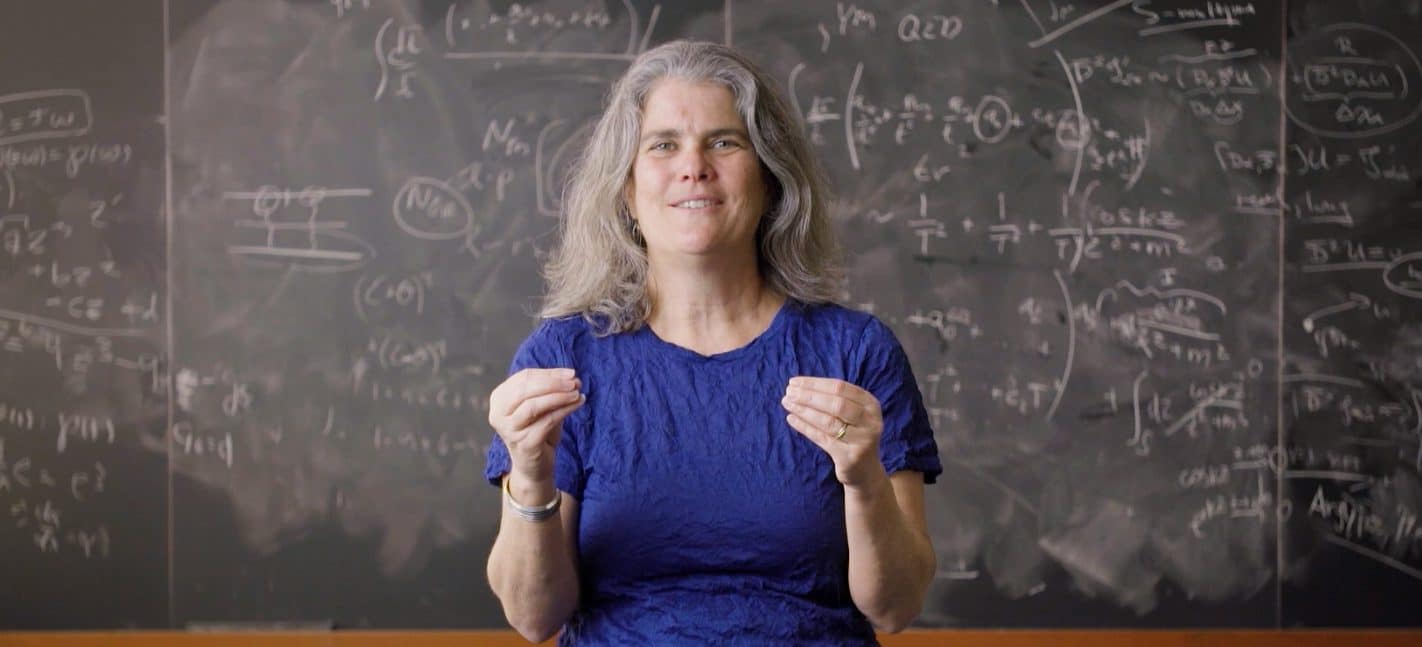 Imagem de Andrea Ghez, astrônoma premiada com o Nobel, com cabelos grisalhos e longos, usando camiseta azul, ao fundo um quadro negro repleto de cálculos astronômicos.