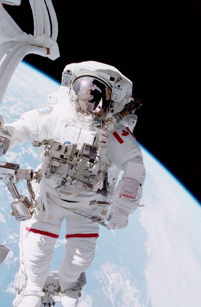 Chris Hadfield, um dos Astronautas Famosos da NASA, flutuando fora da Estação Espacial Internacional (ISS) com traje espacial completo, incluindo capacete e viseira espelhada. Ao fundo, é possível ver a curvatura da Terra, destacando a magnitude da missão e a experiência única de estar no espaço.