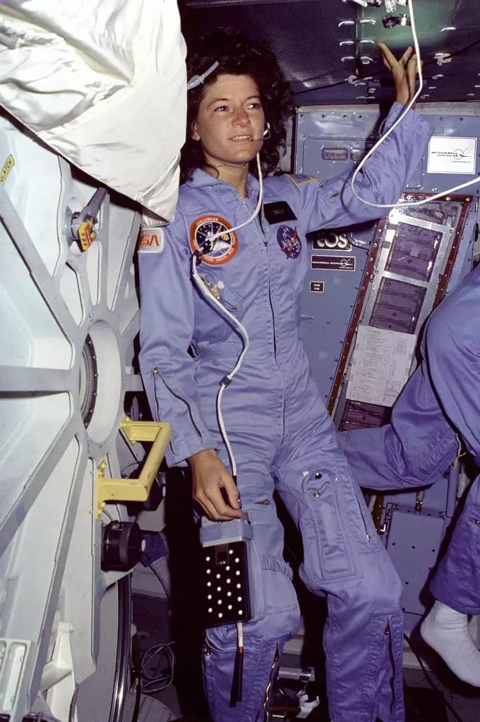 Fotografia histórica de Sally Ride, a primeira mulher americana no espaço, dentro da cabine do ônibus espacial. Ela está com o cabelo volumoso, usando o traje cinza da NASA e fones de ouvido. Ao fundo, é possível ver o painel de controle da nave, destacando a tecnologia da época e a importância da missão espacial por esse feito ela entra na lista de astronautas famosos da nasa
