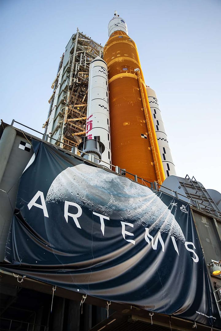 Bandeira do Programa Artemis da NASA em destaque com o imponente foguete SLS ao fundo, representando qual o principal objetivo do Programa Artemis da NASA, que é conduzir missões tripuladas de volta à Lua com tecnologia de ponta.