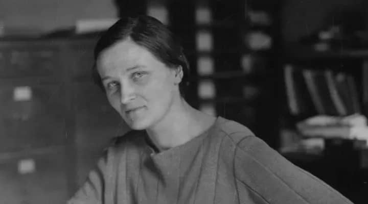 Cecilia Payne-Gaposchkin sentada em uma cadeira, apoiada em uma mesa de escritório, usando roupas da época. A cena é ambientada em uma sala de aparência clássica, com móveis antigos, refletindo o contexto histórico de sua época.