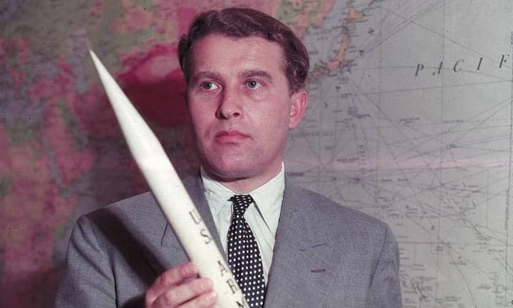 "Wernher von Braun jovem, de olhos claros, vestindo um terno cinza e camiseta branca, segura uma miniatura de foguete com a sigla 'US Army'. Ao fundo, um mapa-múndi destaca sua influência global na engenharia de foguetes.