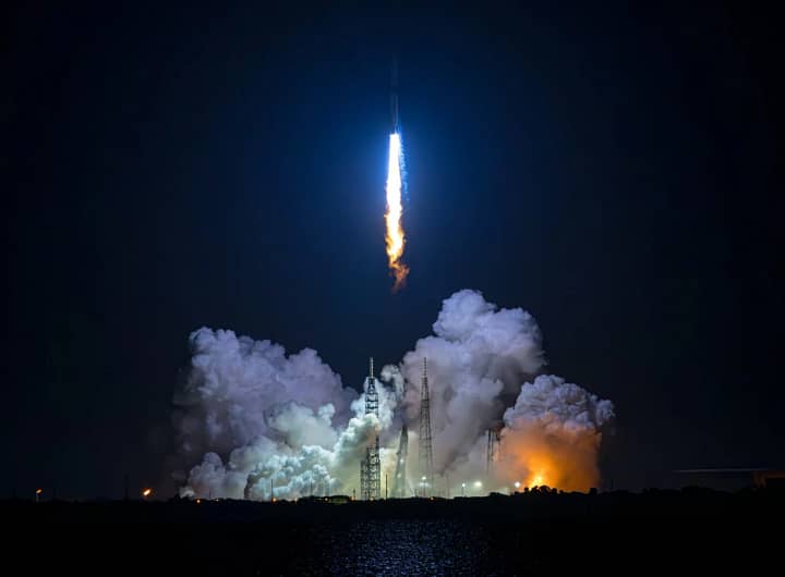 Primeiro voo de teste noturno do foguete New Glenn da Blue Origin, alcançando a órbita em sua tentativa inicial. A imagem captura o foguete saindo do pad de lançamento, com uma enorme coluna de fogo sendo gerada pelos potentes motores. O brilho intenso destaca-se contra o céu escuro da noite.