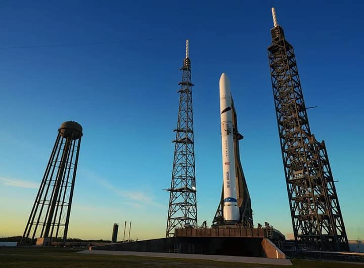 O foguete New Glenn da Blue Origin, de cor branca, está posicionado na plataforma de lançamento durante o dia, aguardando o momento para iniciar seu voo. O céu azul ao fundo contrasta com a estrutura imponente do foguete, pronto para sua missão.