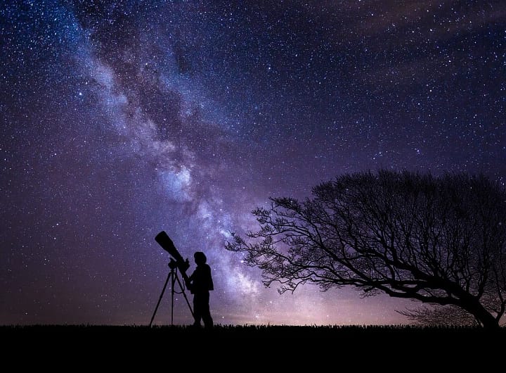Silhueta de uma pessoa com um telescópio apontado para o céu estrelado, com cores roxas e lilás representando uma galáxia distante.