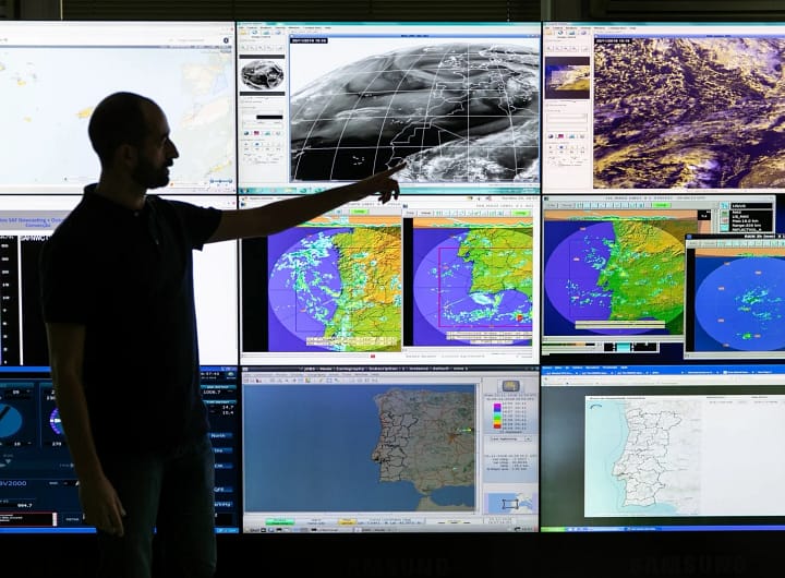 Homem meteorologista em frente a vários monitores exibindo previsões de clima e do tempo, representando o trabalho de monitoramento e análise meteorológica.
