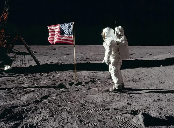 Apollo 11 Buzz Aldrin, o astronauta da missão Apollo 11, saudando a bandeira dos Estados Unidos na superfície da Lua em 1969. Ele está vestido com seu traje espacial, com o visor refletindo a paisagem lunar ao fundo, enquanto a bandeira americana ondula ao vento (apesar da falta de atmosfera) em um gesto simbólico de conquista.