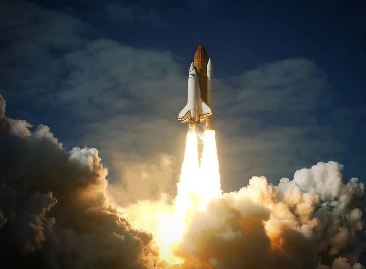 Lançamento de um ônibus espacial, com a enorme potência dos foguetes gerando uma explosão de fogo e fumaça enquanto o veículo ascende em direção ao céu. O ônibus espacial é visto no início de sua trajetória, com a separação dos boosters laterais e o veículo se afastando da base de lançamento. A palavra-chave "Lançamento espacial" é representada pelo evento dinâmico do lançamento.