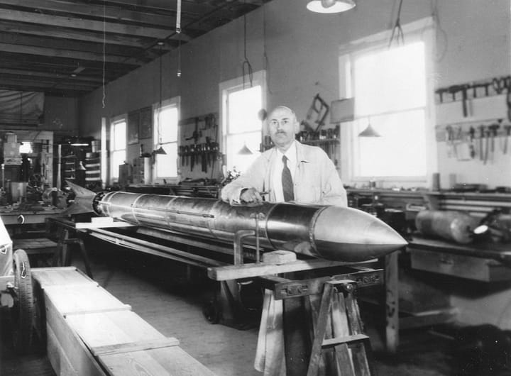 Dr. Robert H. Goddard em frente a um foguete apoiado em uma mesa dentro de um galpão, usando jaleco e gravata. Ao fundo, há ferramentas espalhadas, e o Dr. Goddard tem um olhar sério, focado em seu trabalho