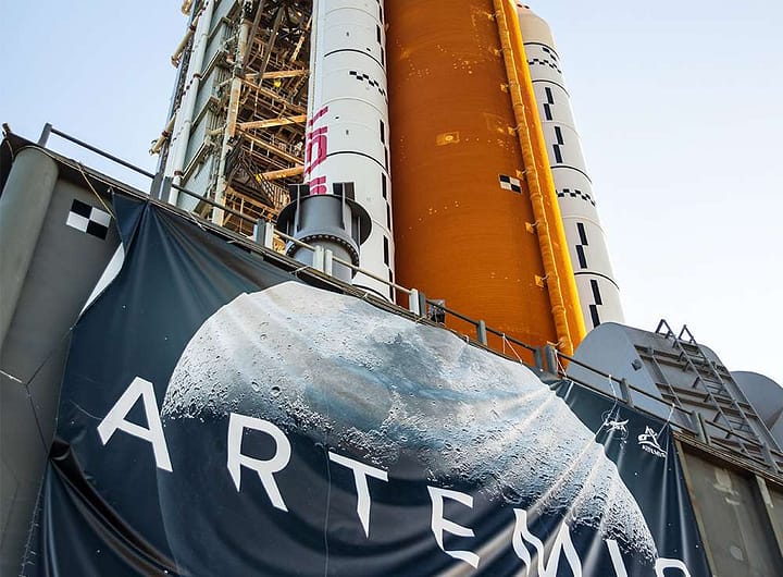 Bandeira do Programa Artemis da NASA em destaque com o imponente foguete SLS ao fundo, representando qual o principal objetivo do Programa Artemis da NASA, que é conduzir missões tripuladas de volta à Lua com tecnologia de ponta.