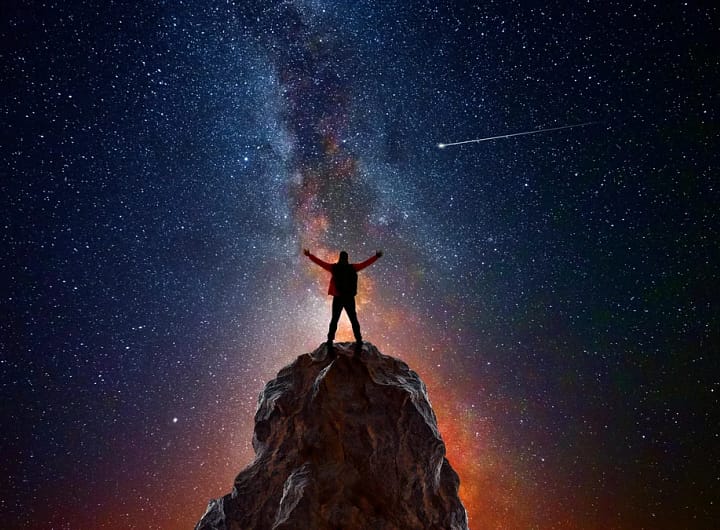 Pessoa no topo de uma montanha com os braços levantados em direção ao céu noturno estrelado, observando um cometa e o braço da Via Láctea, representando como a astronomia define a conexão humana com o universo.