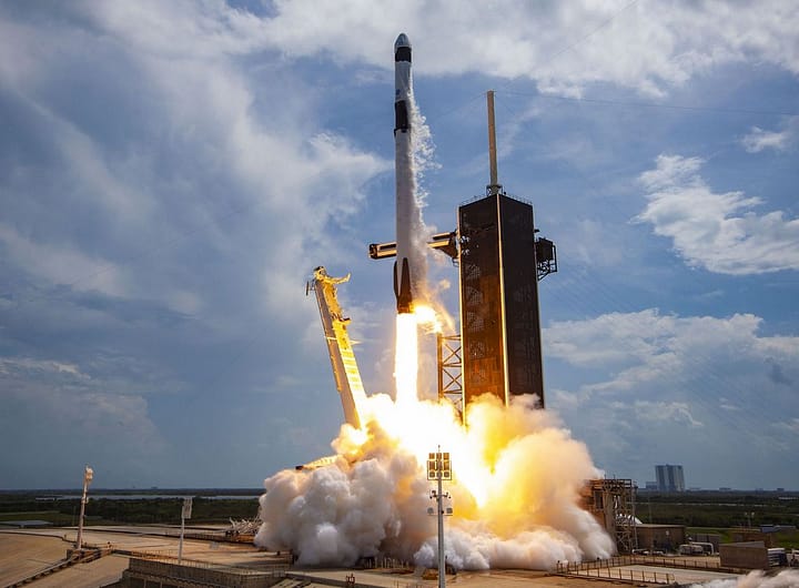Foguete Falcon 9 sendo lançado da base, com fumaça e chamas saindo dos motores enquanto sobe em direção ao céu azul.
