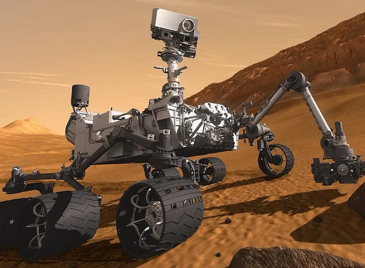 Representação artística do rover Curiosity da NASA em Marte, mostrando o veículo robótico de seis rodas sobre uma superfície rochosa avermelhada, com colinas e o céu marciano em tons alaranjados ao fundo. O rover aparece equipado com seu mastro de câmeras levantado, seus painéis e instrumentos cobertos por poeira marciana, enquanto realiza exploração científica da paisagem ao redor.