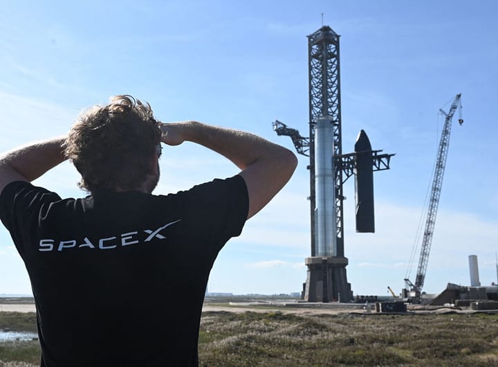Captura do Super Shuper heavy Elon Musk de costas, vestindo uma camiseta preta com o logo da SpaceX, observa o Super Heavy e a Starship sendo segurados pelos chopsticks do Mechazilla.
