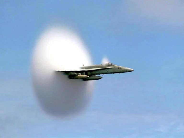 Caça supersônico em plena aceleração, atravessando a barreira do som e gerando uma onda de choque visível. O avião é de cor cinza metálico e o céu está claro e sem nuvens.