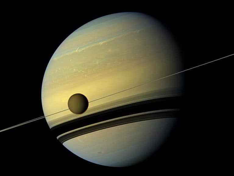 lua Titã em primeiro plano, envolta por uma névoa alaranjada, com Saturno e seus anéis visíveis ao fundo no espaço profundo.