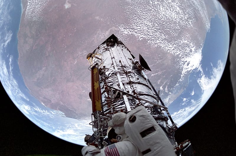 Visão a bordo do ônibus espacial STS-61 do Telescópio Espacial Hubble Com o planeta Terra ao fundo, o Telescópio Espacial Hubble é mostrado exibindo painéis solares novos e modificados, acondicionados na lateral. Um astronauta em caminhada espacial é visível em primeiro plano na imagem. O 59º voo do ônibus espacial e o último de 1993, a STS-61 foi uma das missões tripuladas mais desafiadoras e complexas já realizadas. Durante um recorde de cinco caminhadas espaciais consecutivas, totalizando 35 horas e 28 minutos, duas equipes de astronautas concluíram a primeira manutenção do Hubble desde seu lançamento em abril de 1990. A missão STS-61 foi lançada a bordo do Ônibus Espacial Endeavour em 2 de dezembro de 1993