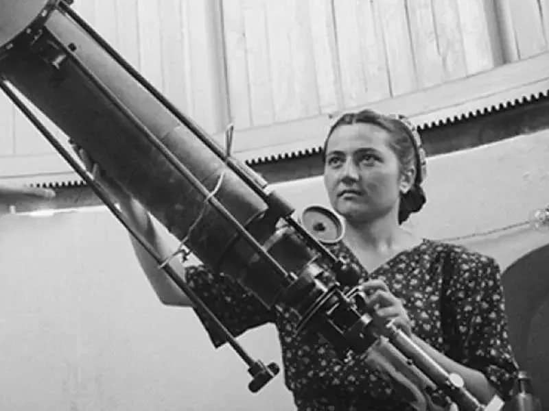 Cecilia Payne-Gaposchkin manipulando um telescópio grande, com foco no instrumento enquanto observa o céu. Ela está em um ambiente de pesquisa astronômica, demonstrando seu trabalho na área de estudos astronômicos.