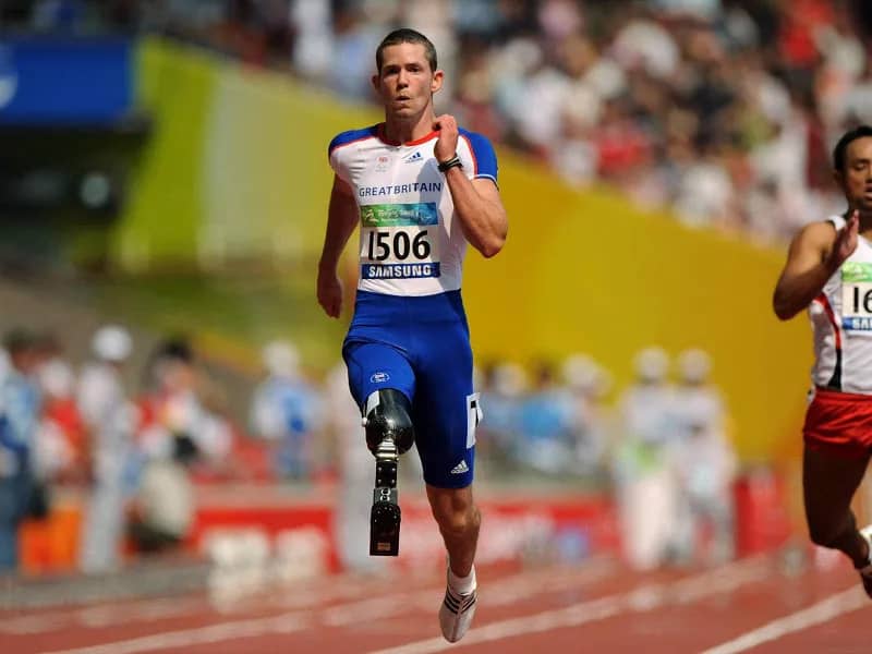 John McFall competindo nas Paralimpíadas, usando sua prótese de perna durante uma prova de atletismo. Ele está em plena ação, com sua postura focada e determinada enquanto corre na pista, vestindo o uniforme Paralímpico e os tênis especiais para atletas com deficiência. A imagem transmite sua força, habilidade e dedicação ao esporte, representando o espírito de superação das Paralimpíadas.